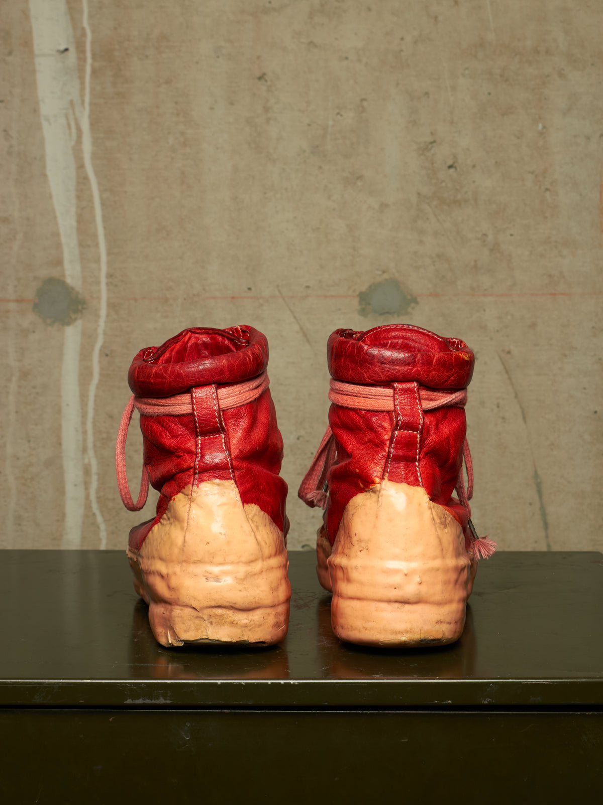 A pair of red leather high-top boots with chunky beige platform soles, shown from the back on a dark surface against a textured concrete wall.