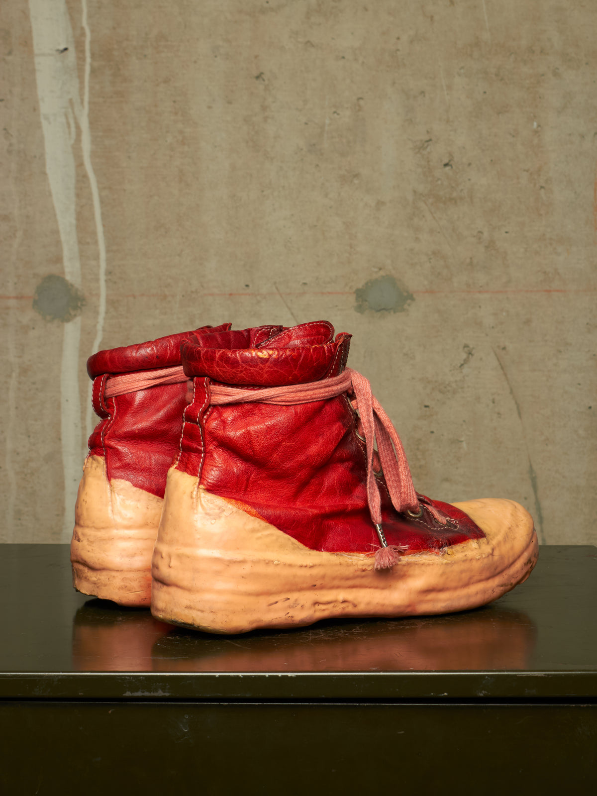 A pair of vintage red leather high-top work boots with tan rubber soles, displayed on a dark reflective surface against a textured concrete wall.