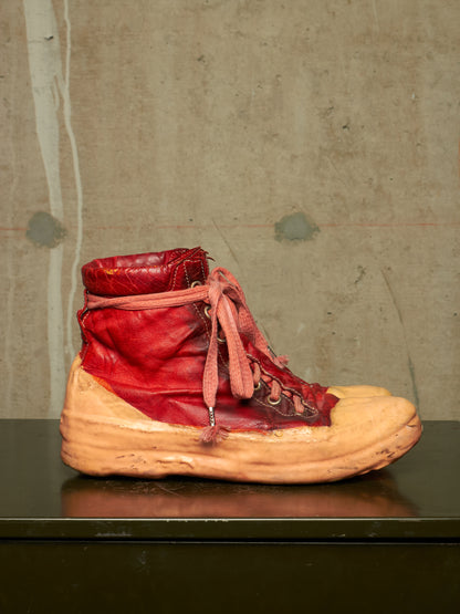 A single high-top boot with a glossy red leather upper, dusty pink laces, and a thick, distressed tan sole, displayed on a dark surface against a textured concrete wall.