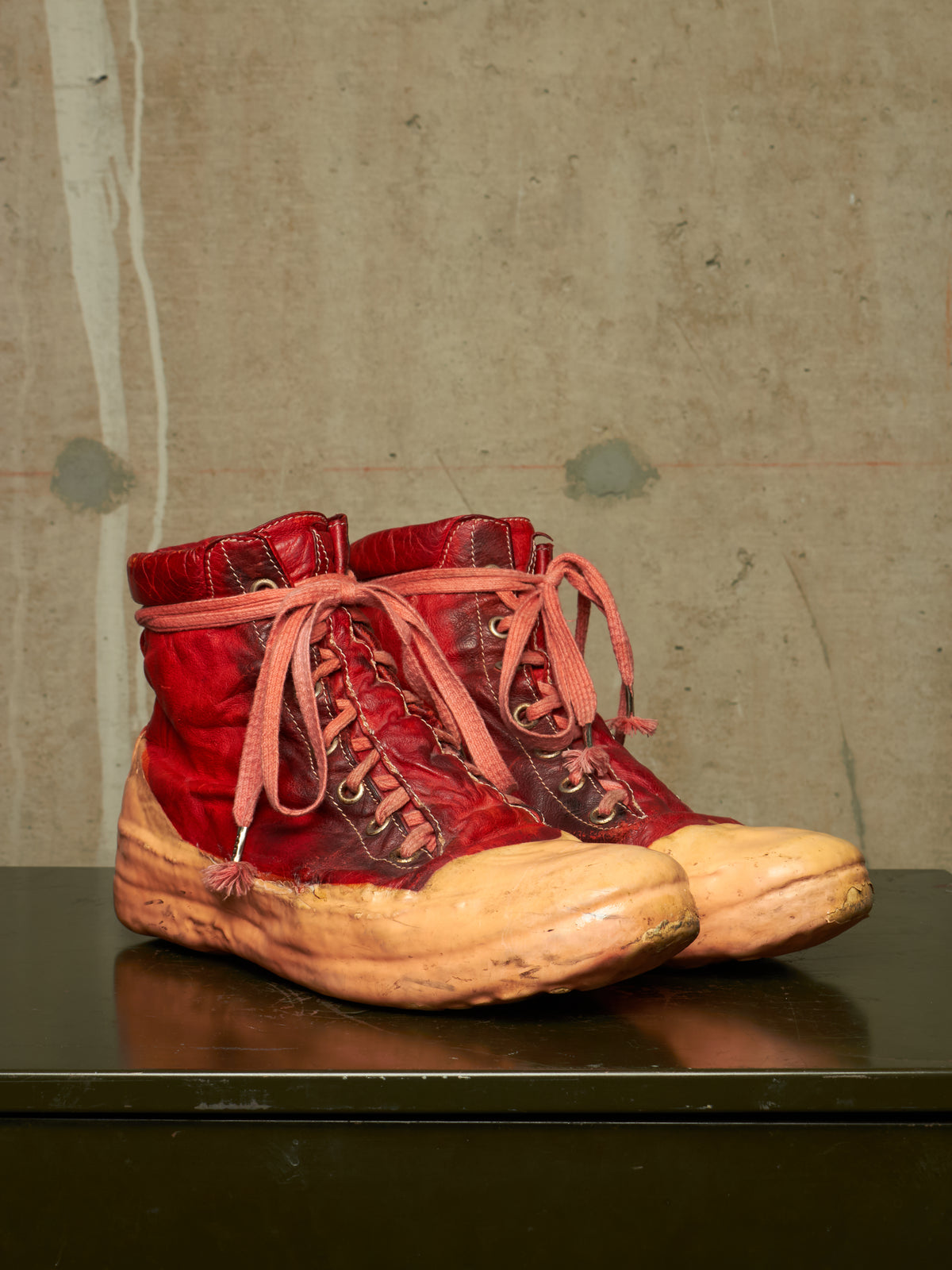 A pair of distressed red high-top lace-up boots with thick tan soles, displayed on a dark reflective surface against a textured concrete wall.
