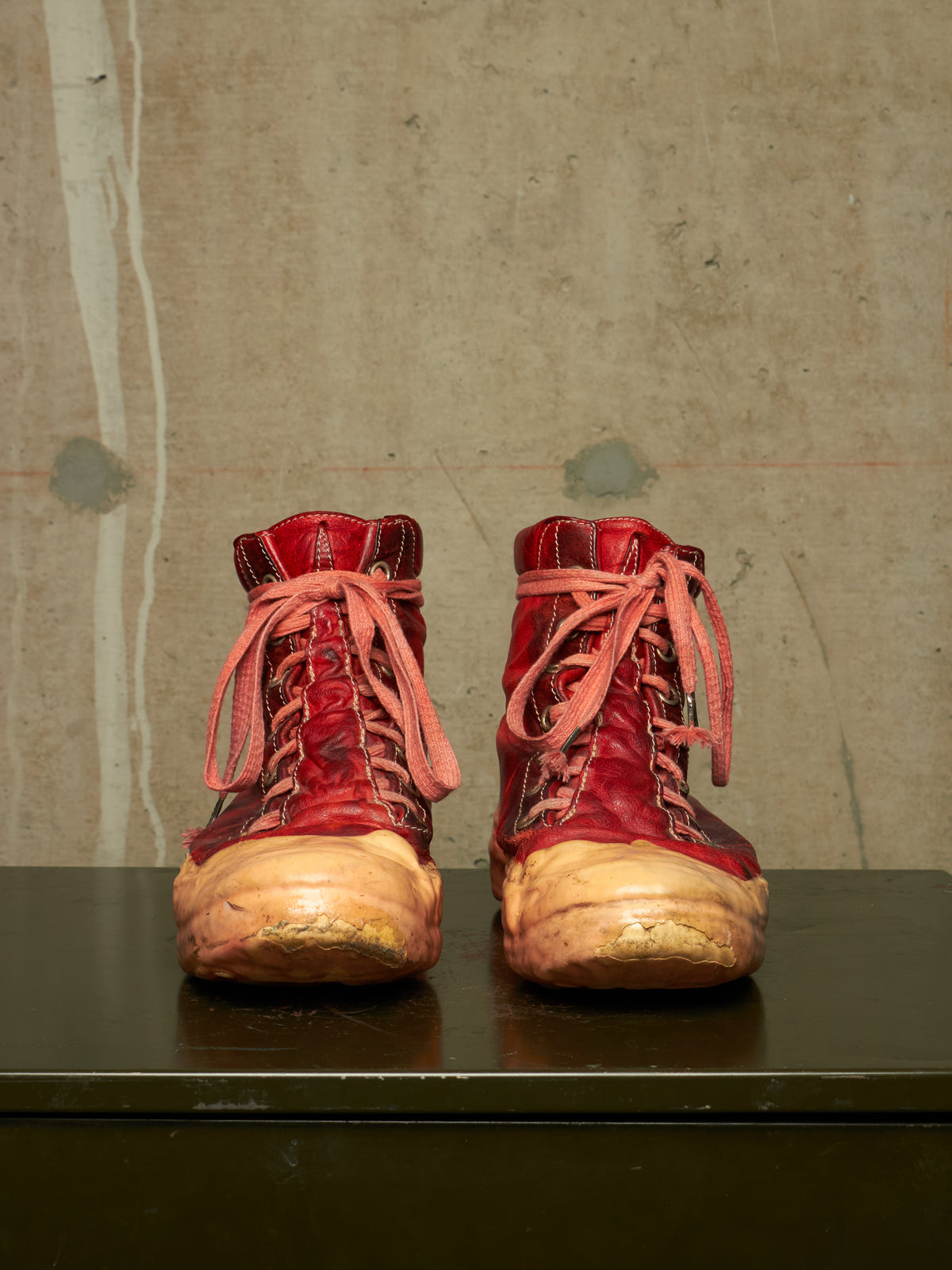 A pair of distressed red leather lace-up boots with heavily worn tan soles, displayed side-by-side on a dark surface against a textured concrete wall.