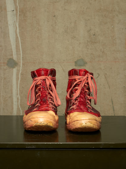 A pair of distressed red leather lace-up boots with heavily worn tan soles, displayed side-by-side on a dark surface against a textured concrete wall.