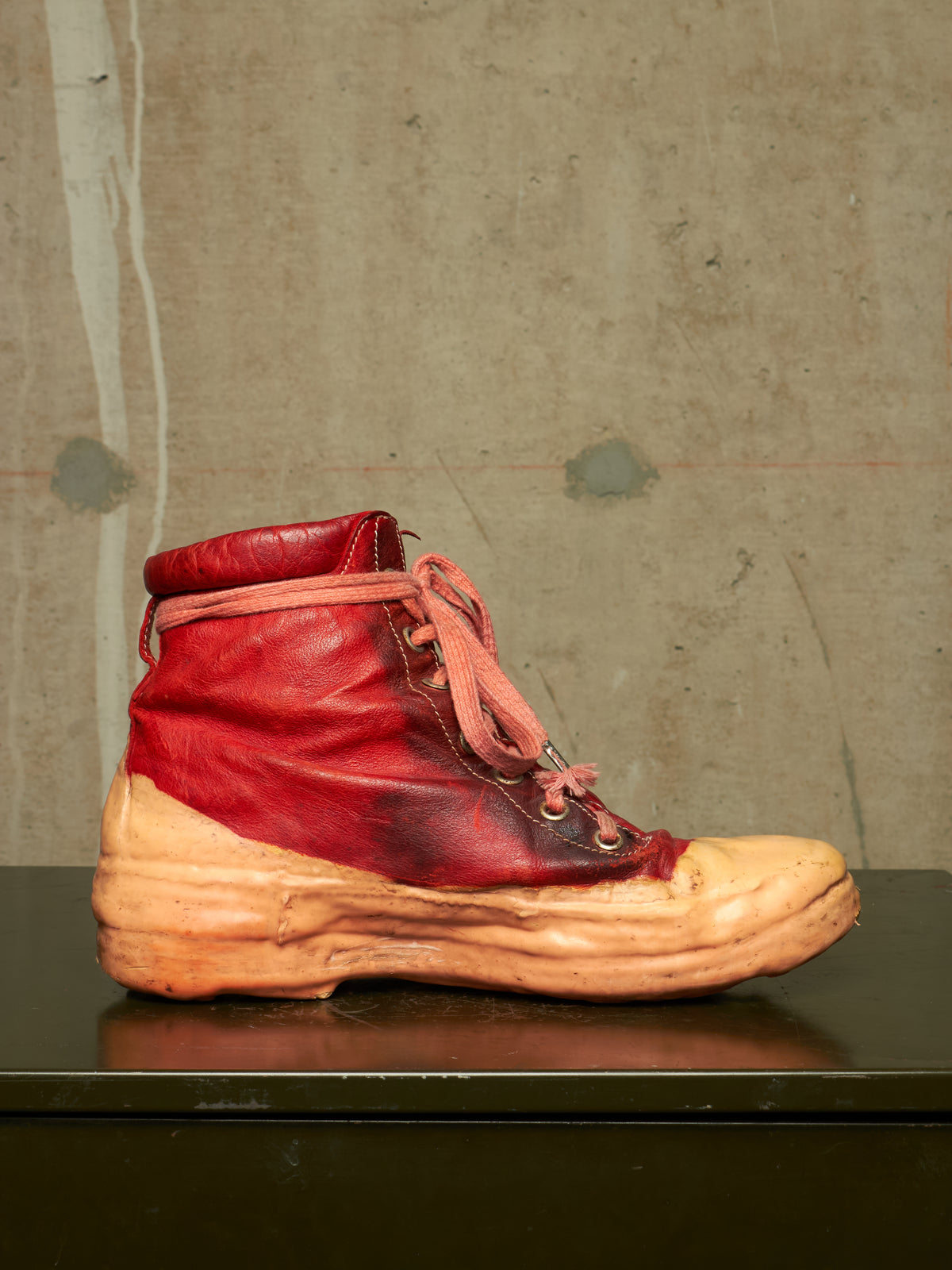 A single high-top leather boot with a distressed red upper and a thick tan sole, displayed on a dark surface against a textured concrete wall.