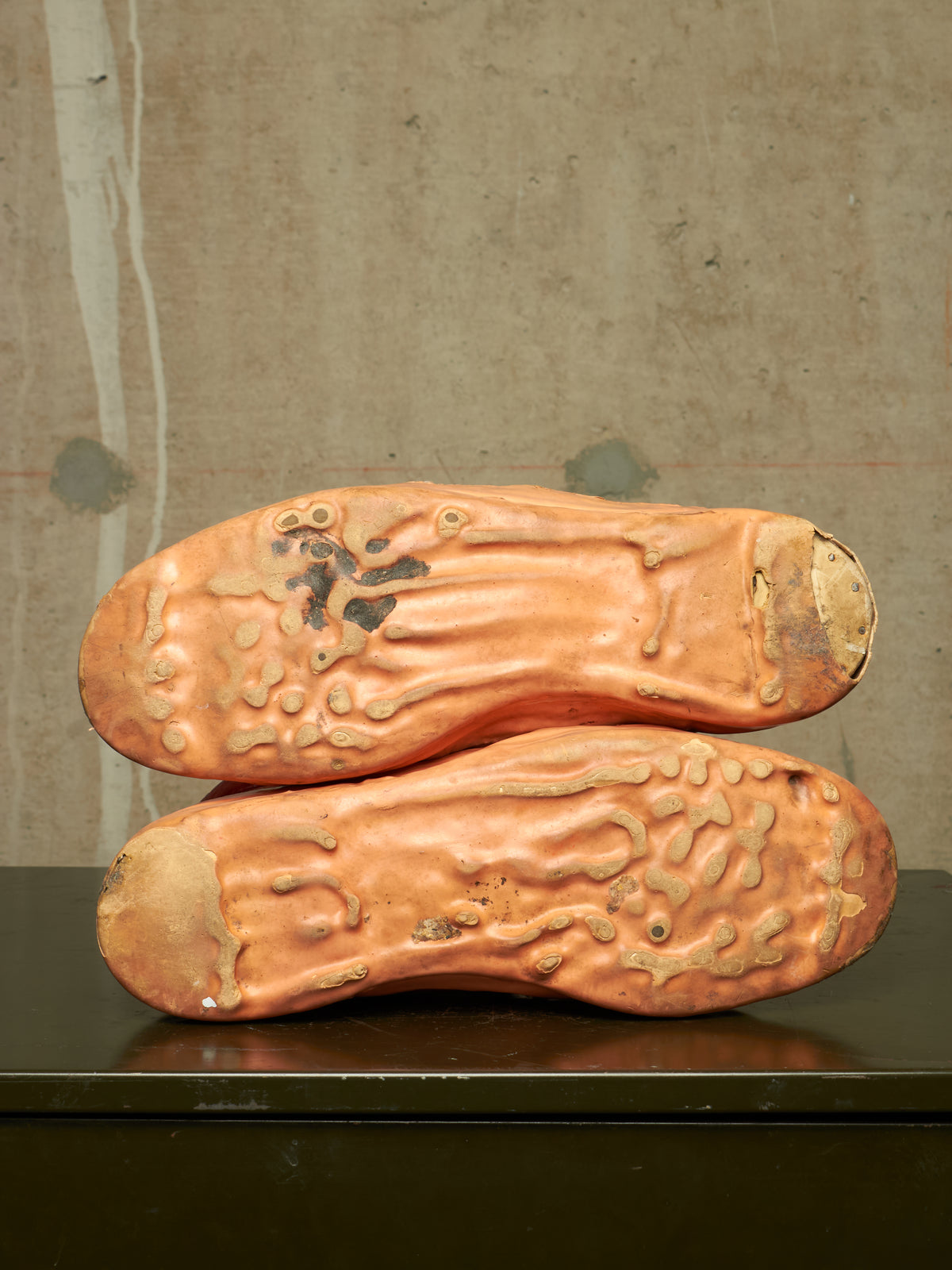 A pair of vintage leather shoes stacked on top of each other, showing their heavily textured, distressed soles in a warm terracotta orange-brown color against a neutral concrete wall background.