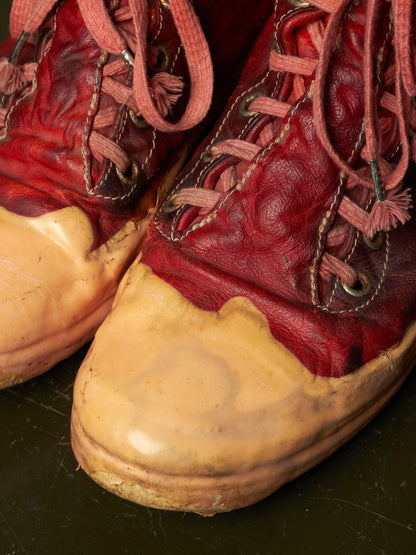 A close-up of a pair of worn red leather work boots with thick tan rubber toe caps and dusty pink laces, resting on a dark surface.