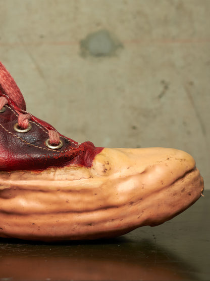 A close-up of a unique high-top sneaker featuring a thick, sculptural layered tan sole and a dark red leather lace-up upper with metal eyelets, set against a textured concrete background.