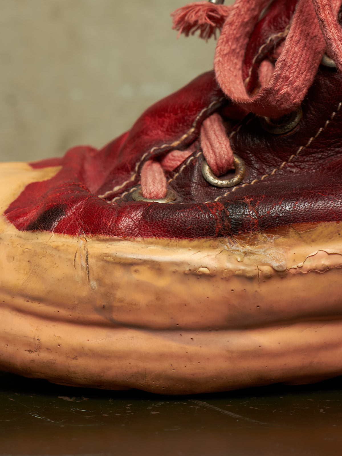 A close-up of a vintage, heavily worn leather shoe with deep red and dark brown leather upper, dusty red laces, and a thick, weathered tan rubber sole.