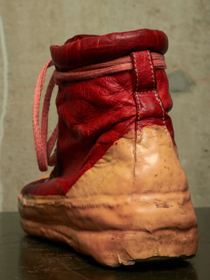 A single high-top boot shown from the back, featuring a red leather upper with pink laces and a heavily worn, tan sole.