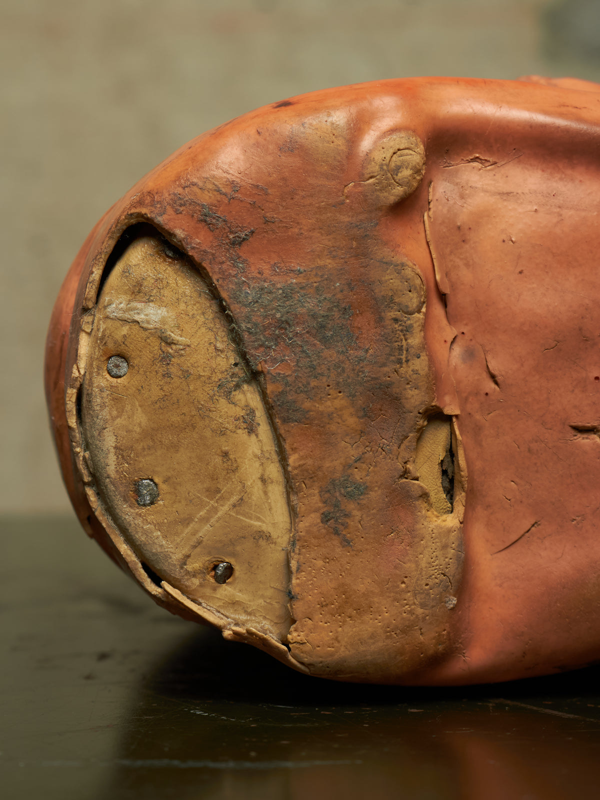 Close-up of the sole and heel of a worn terracotta-colored shoe or boot, showing peeling reddish-brown outer material and a weathered tan sole with metal rivets, resting on a dark surface.