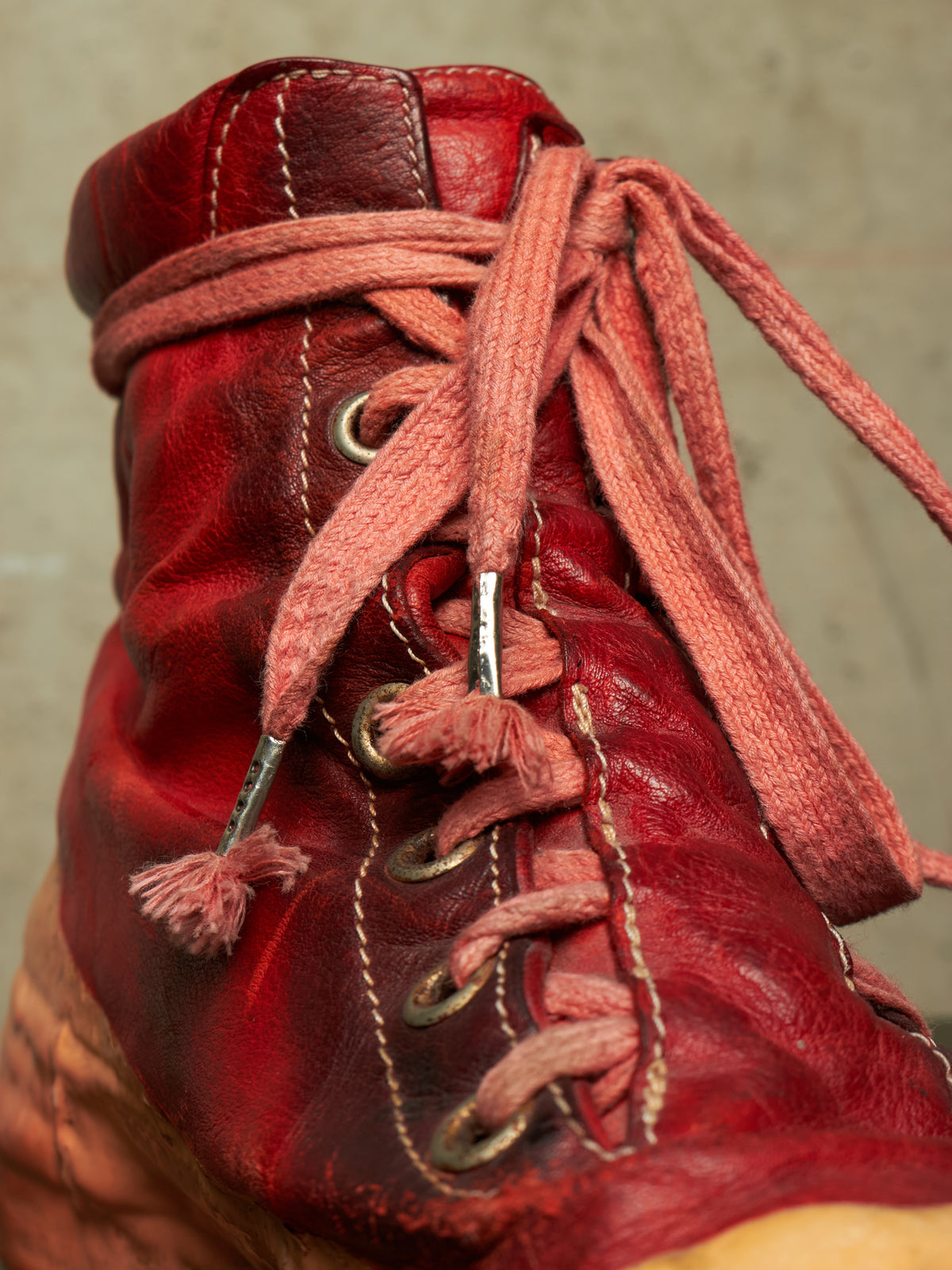 A close-up of a distressed red leather lace-up boot with matching red laces and metal eyelets, set against a neutral textured background.