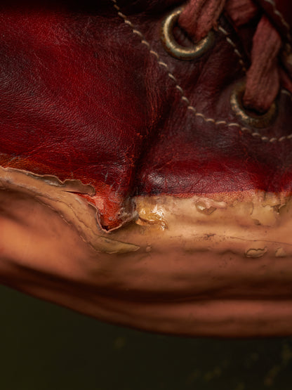 Extreme close-up of a worn dark red leather shoe or boot sole with visible peeling layers, tan wear marks, and part of a brown laces eyelet area.