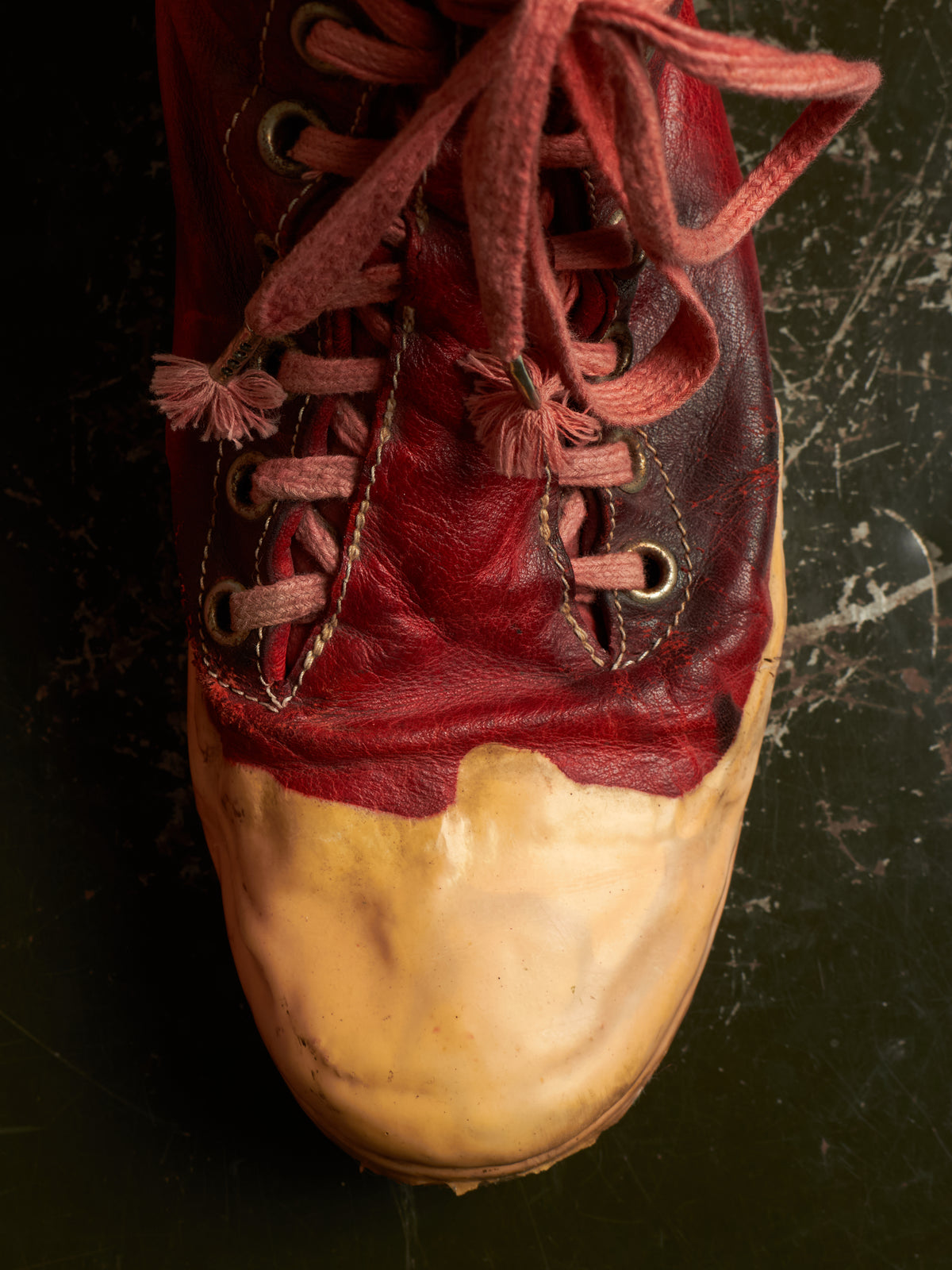 A close-up of a single leather shoe with a dark red upper, matching laces, and a heavily worn tan rubber toe cap, set against a dark textured background.