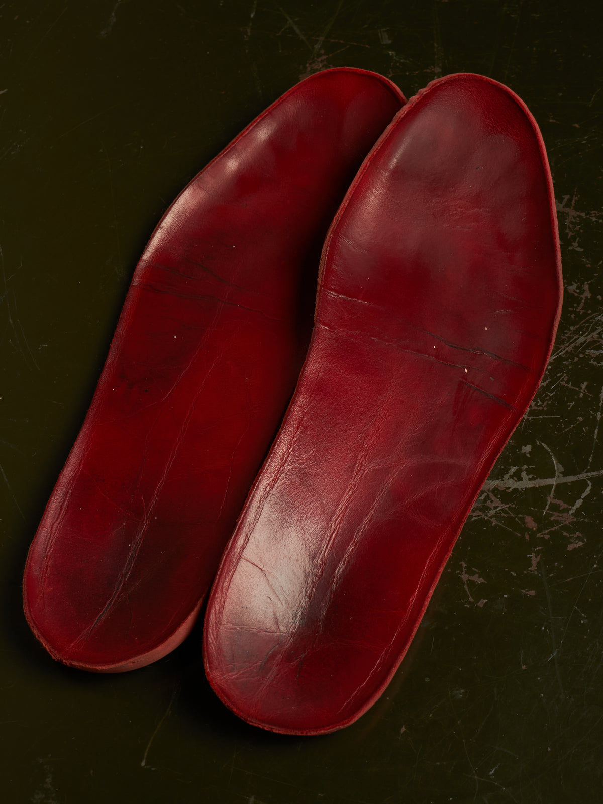 A pair of red leather shoe insoles with a glossy finish, laid flat on a dark textured background.