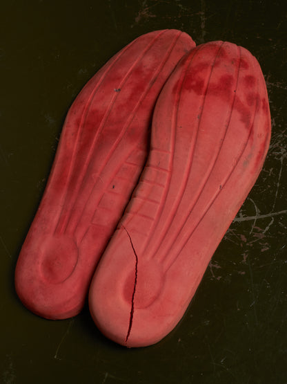A pair of red molded shoe insoles or footbeds laid flat on a dark textured surface, showing the underside with molded ridges and a visible crack on one piece.
