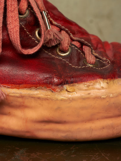 A close-up of a red leather sneaker with matching red laces and a thick tan sole, photographed against a neutral beige background.