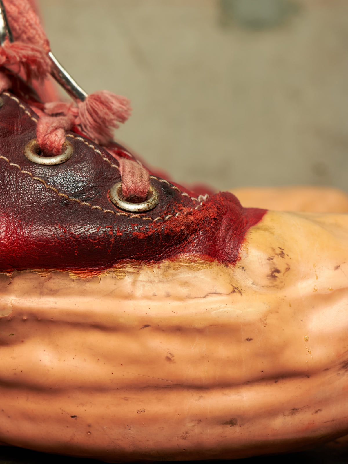 A close-up of a leather shoe or boot with dark reddish-brown leather, pinkish-red laces, and a beige/tan textured sole or base against a blurred gray background.