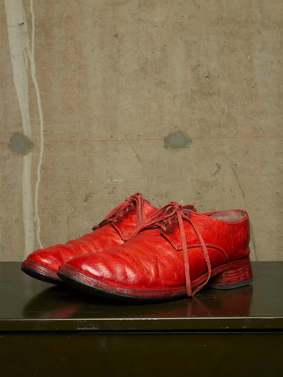 A pair of bright red leather lace-up dress shoes with a textured, embossed pattern, displayed on a dark reflective surface against a textured concrete wall.