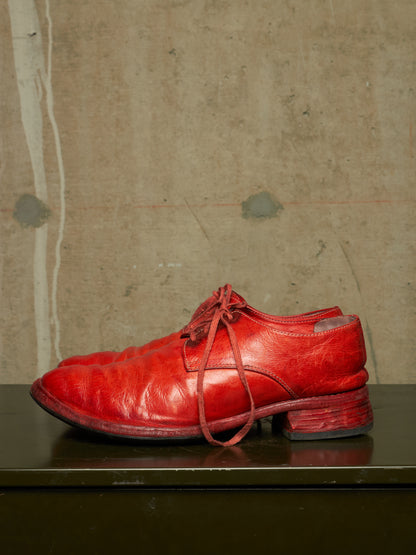 A pair of bright red leather lace-up dress shoes with a distressed, cracked texture, resting on a dark reflective surface against a textured concrete wall.