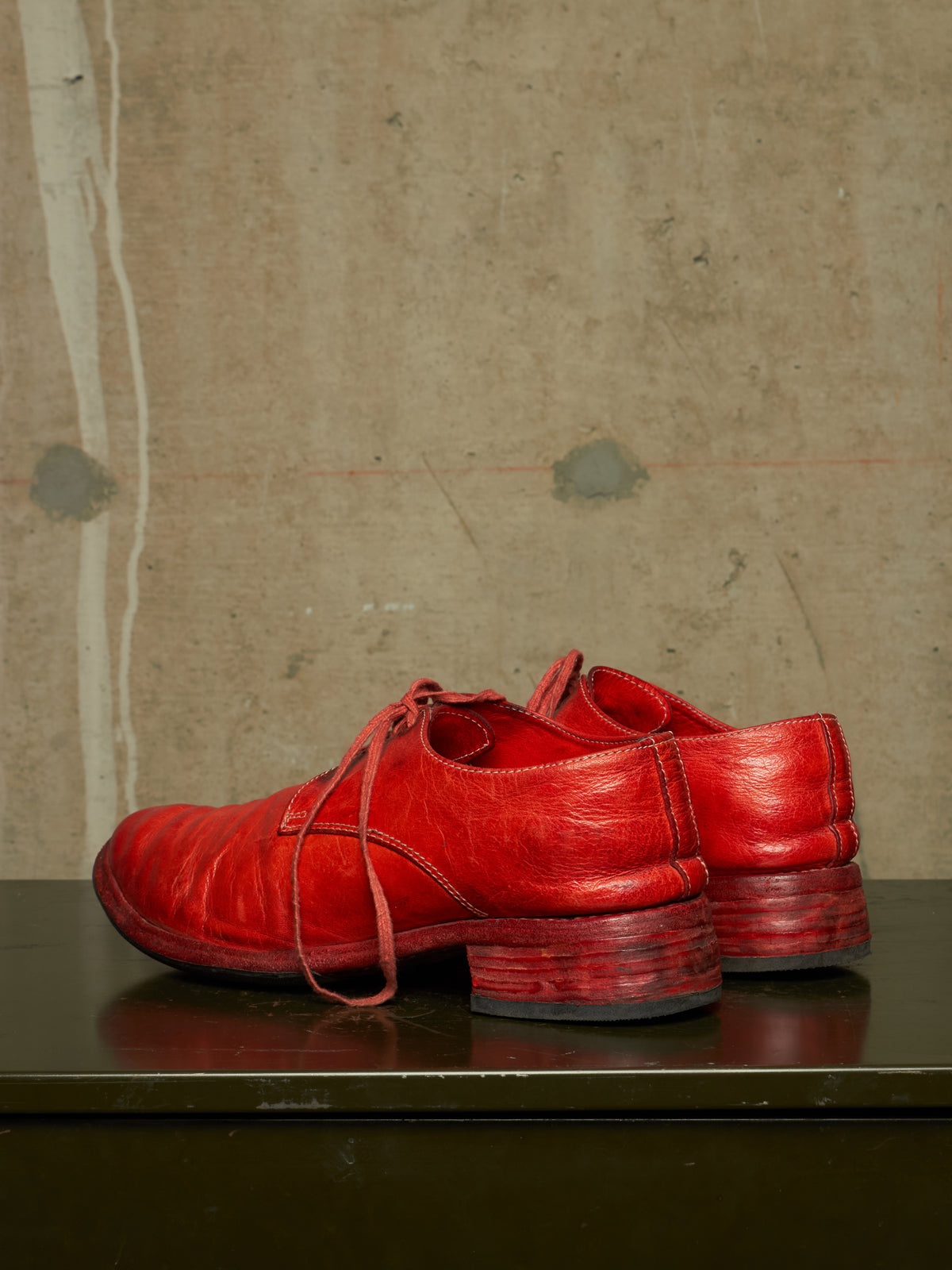 A pair of bright red leather lace-up shoes with stacked block heels, displayed on a dark reflective surface against a textured concrete wall.
