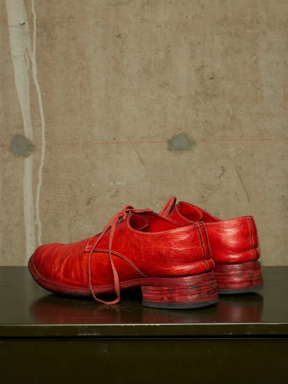 A pair of bright red leather lace-up shoes with stacked block heels, displayed on a dark reflective surface against a textured concrete wall.