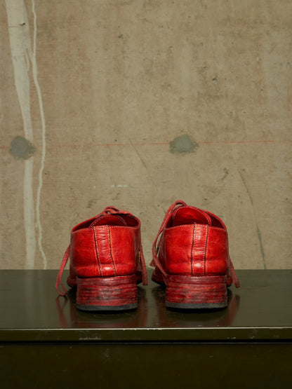 A pair of distressed red leather lace-up shoes shown from the back, resting on a dark reflective surface against a textured concrete wall.