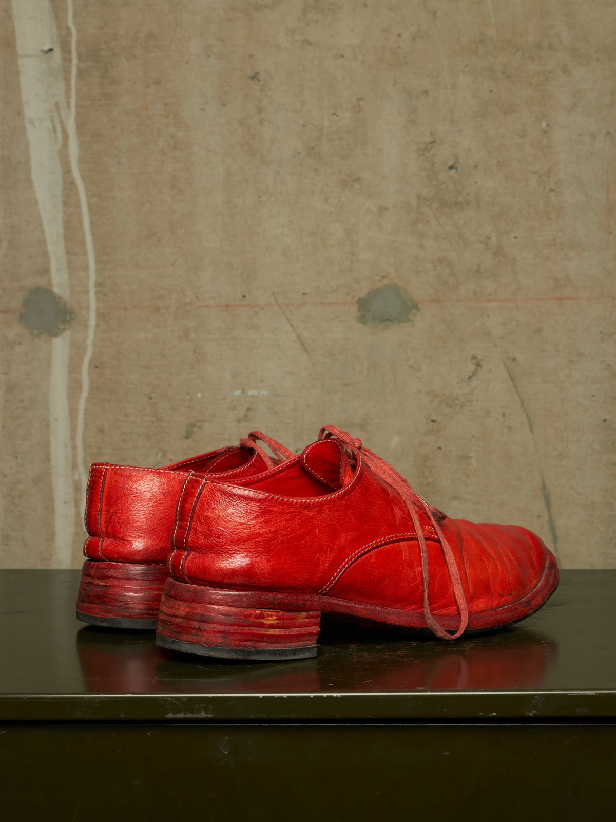 A pair of distressed red leather lace-up shoes with stacked block heels, displayed on a dark reflective surface against a textured concrete wall.