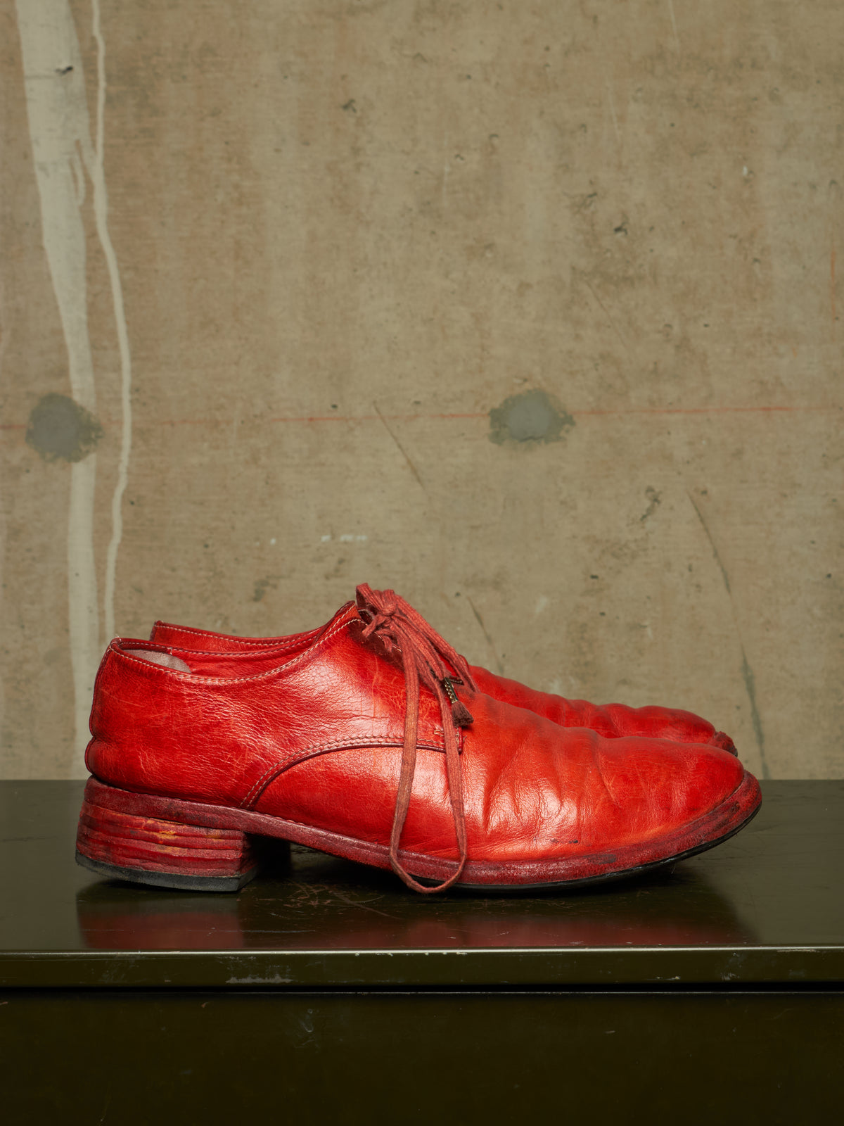 A pair of bright red leather lace-up shoes with low block heels, displayed on a dark reflective surface against a textured concrete wall.