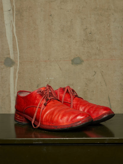 A pair of bright red leather lace-up shoes with a distressed finish, displayed on a dark reflective surface against a textured concrete wall.