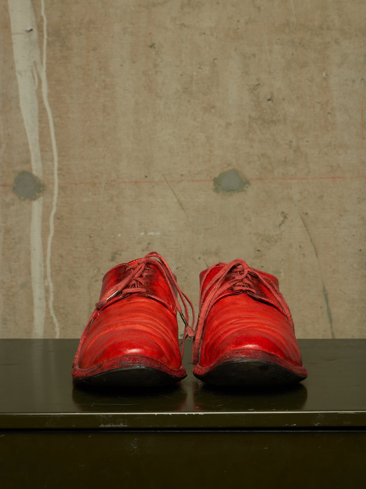 A pair of bright red leather lace-up shoes with a glossy finish, displayed side-by-side on a dark green surface against a textured concrete wall.