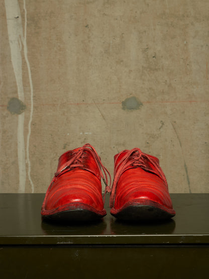 A pair of bright red leather lace-up shoes with a glossy finish, displayed side-by-side on a dark green surface against a textured concrete wall.