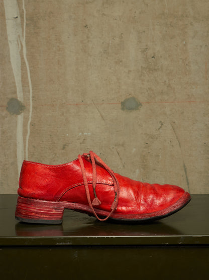 A single red leather lace-up shoe with a chunky stacked heel, resting on a dark reflective surface against a textured concrete wall.