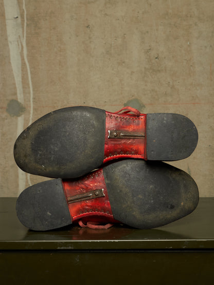 A pair of red leather boots shown from the sole side, resting on a dark green surface against a textured beige wall.