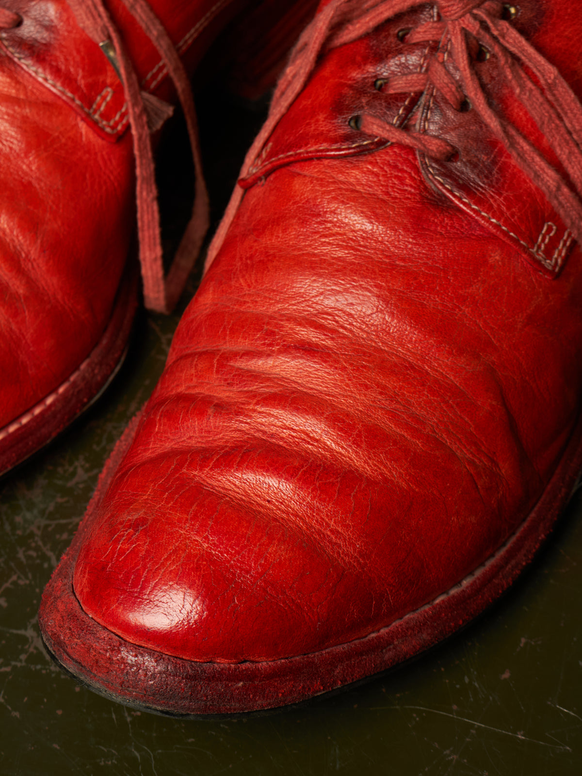A close-up shot of a pair of distressed red leather lace-up shoes resting on a dark, textured surface.