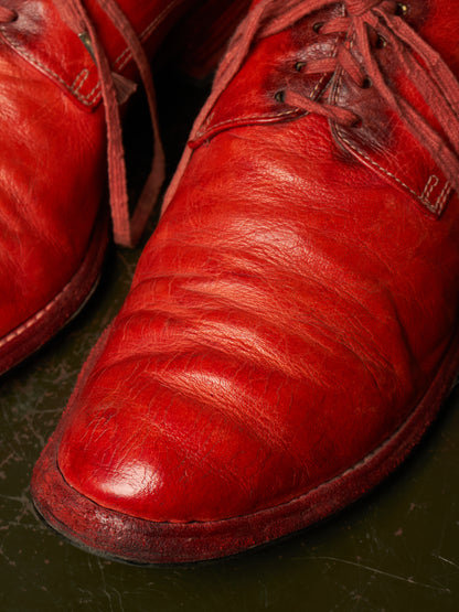 A close-up shot of a pair of distressed red leather lace-up shoes resting on a dark, textured surface.