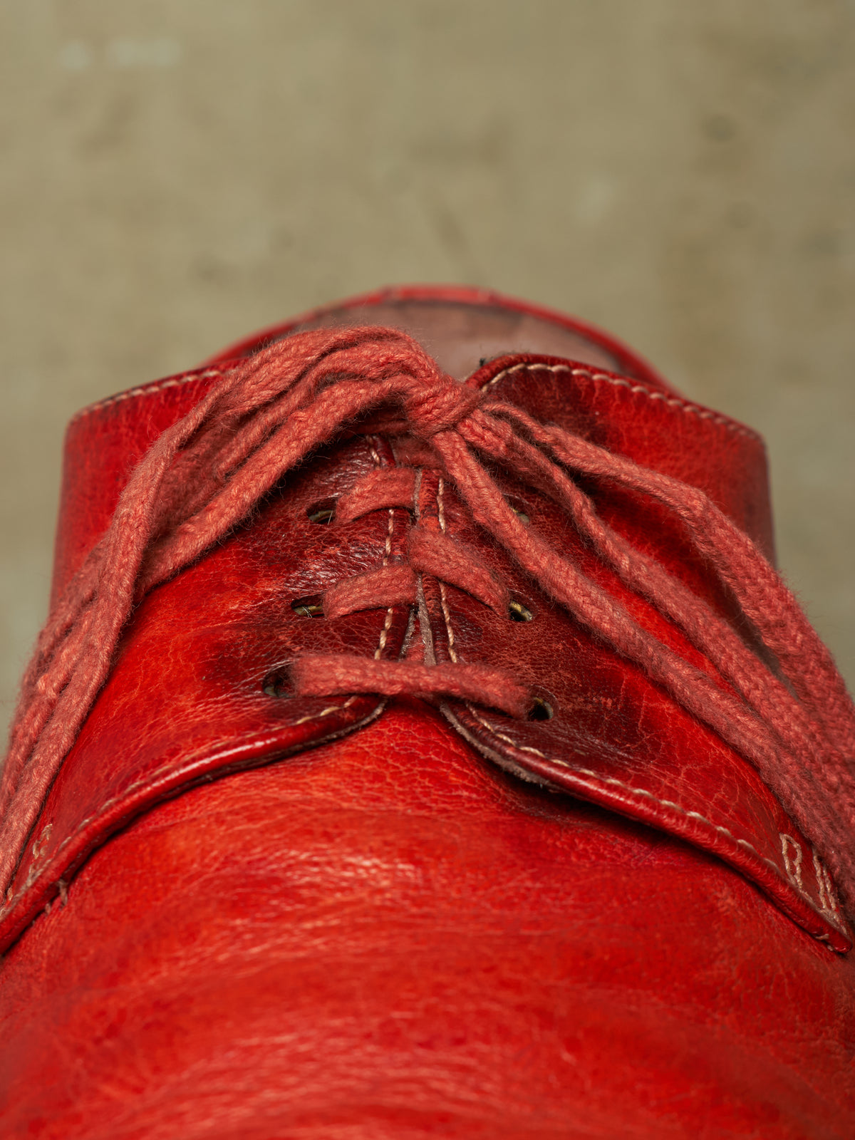 A close-up of a red leather lace-up shoe, focusing on the textured leather and the tied laces against a neutral gray background.