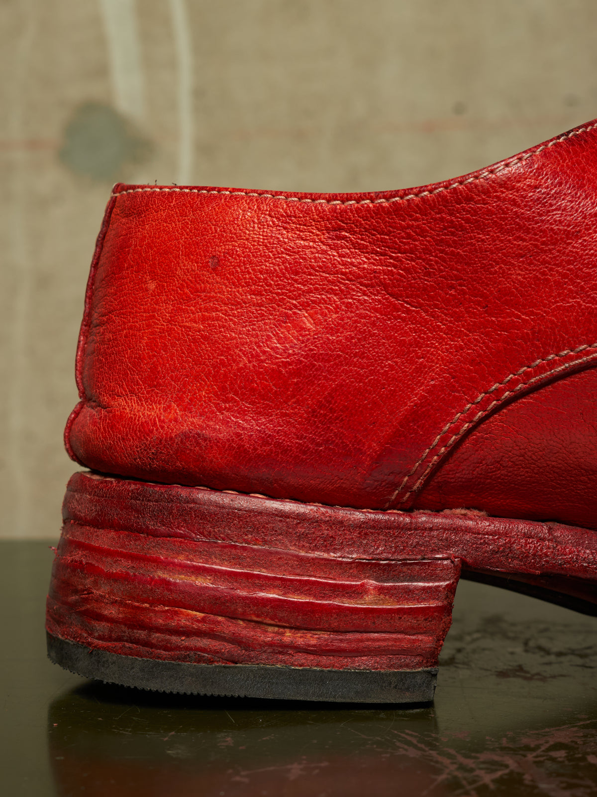 A close-up shot of a distressed red leather shoe heel, showing the textured leather, stitching, and a chunky stacked red block heel on a dark reflective surface.