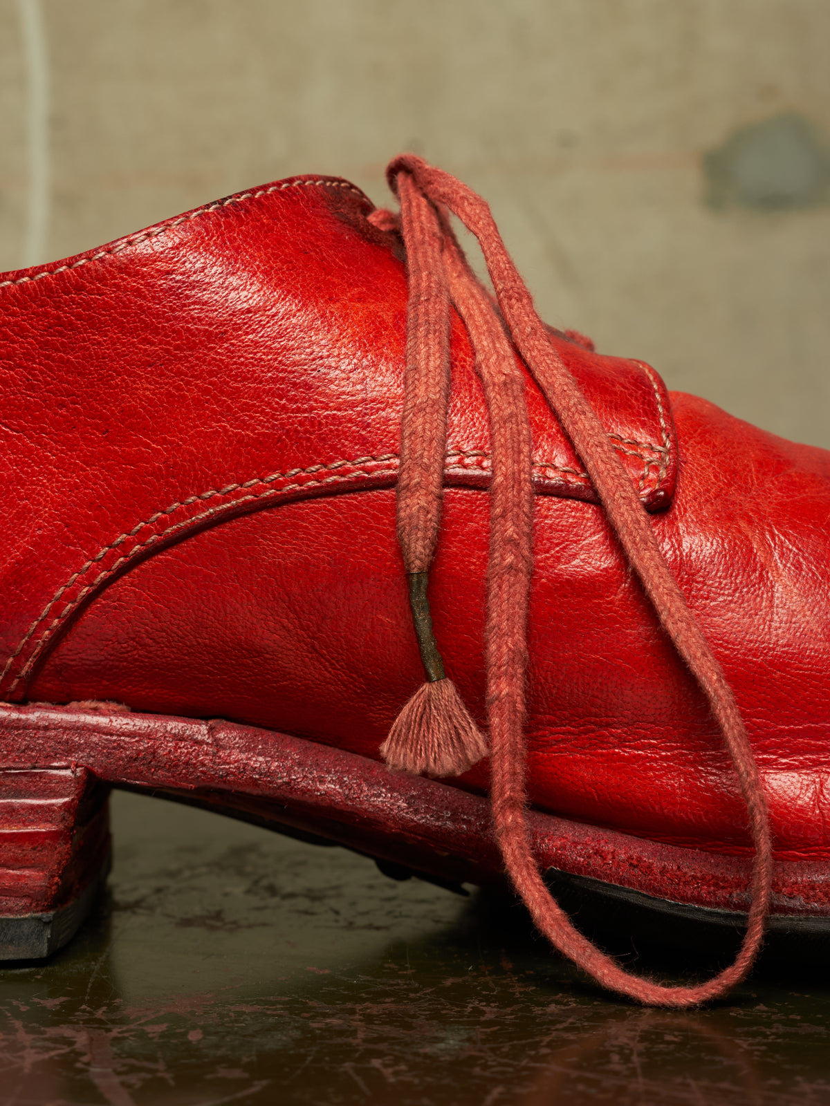A close-up of a red leather boot with matching red laces and a low block heel, resting on a dark reflective surface against a textured gray background.