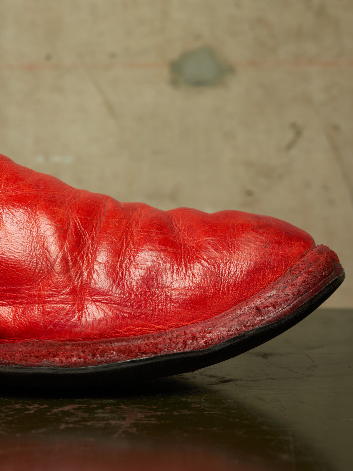 A close-up of a bright red leather boot showing the textured toe and sole edge, resting on a dark reflective surface against a mottled beige background.