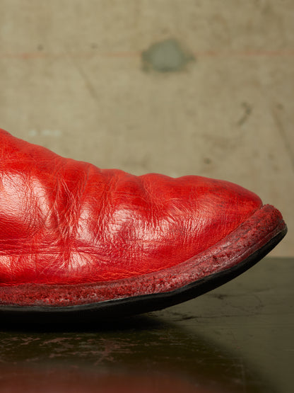 A close-up of a bright red leather boot showing the textured toe and sole edge, resting on a dark reflective surface against a mottled beige background.