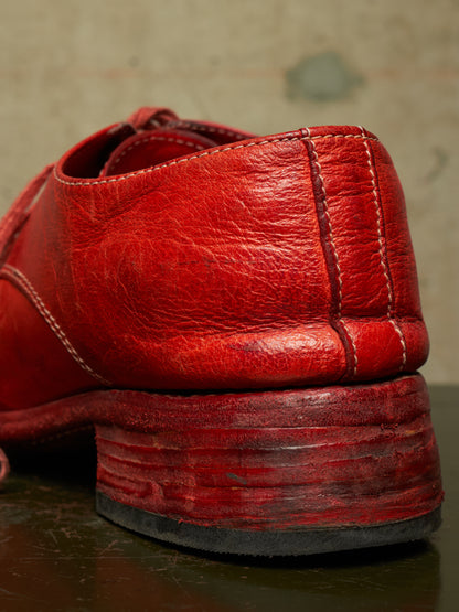 A close-up shot of a distressed red leather shoe heel, showing the textured leather, contrast stitching, and stacked block heel on a dark reflective surface.