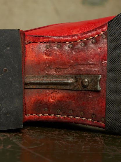 A close-up of a red leather weightlifting belt with a metal toggle clasp and black fabric side panels, resting on a dark surface.