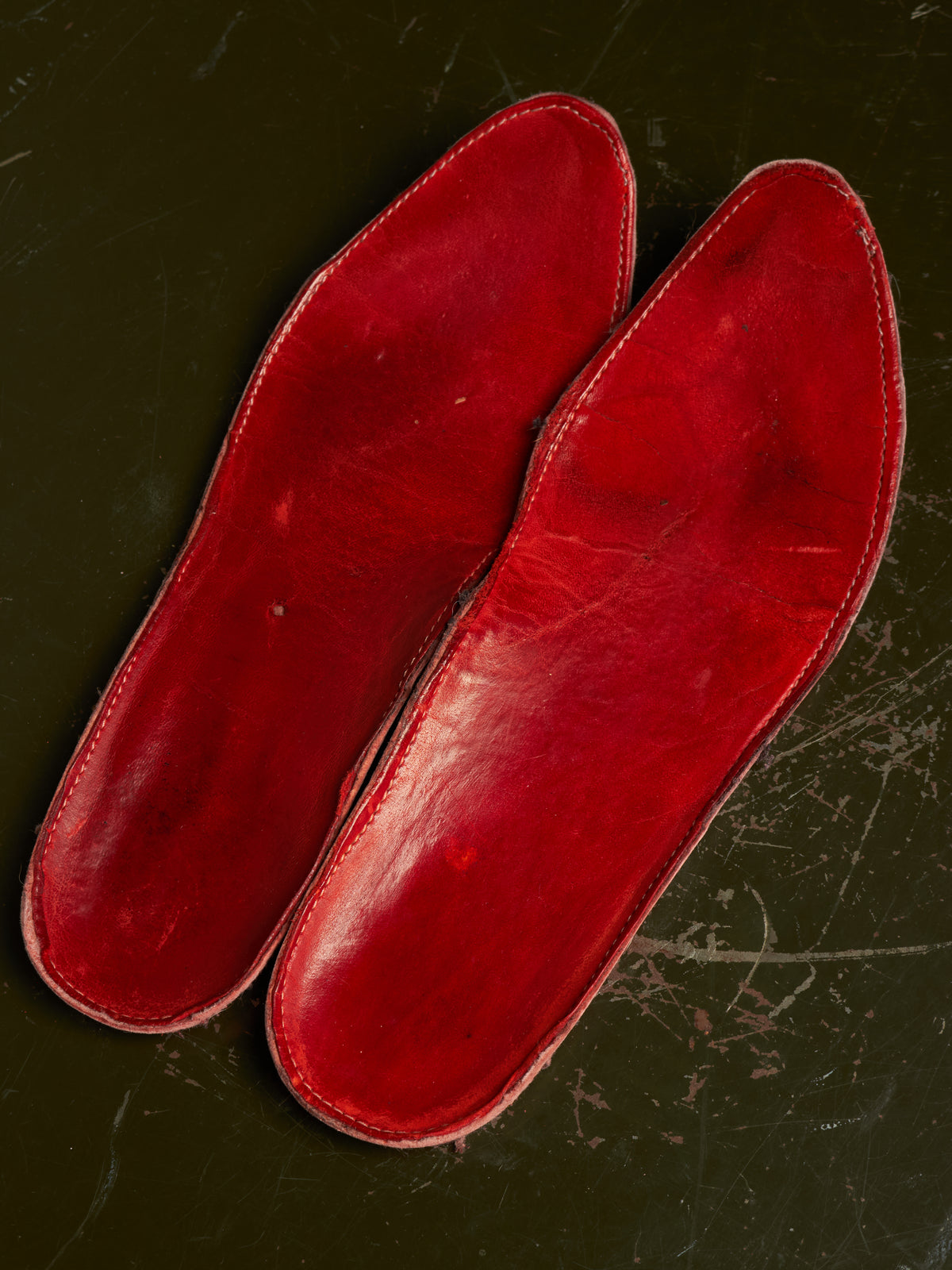 A pair of red leather shoe insoles or footbeds laid flat on a dark, scratched surface.
