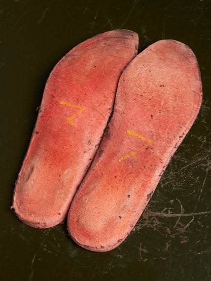 Two pinkish-red insole or footbed pieces placed on a dark scratched surface, with small yellow markings on each piece.