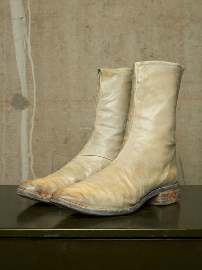 A pair of worn beige leather cowboy boots with pointed toes and dark soles, resting on a dark surface against a textured concrete wall.