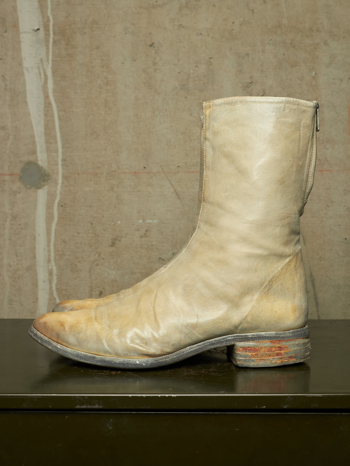 A single light beige leather boot with a distressed, weathered finish, featuring a low stacked heel and a zip closure on the side, resting on a dark surface against a textured concrete wall.