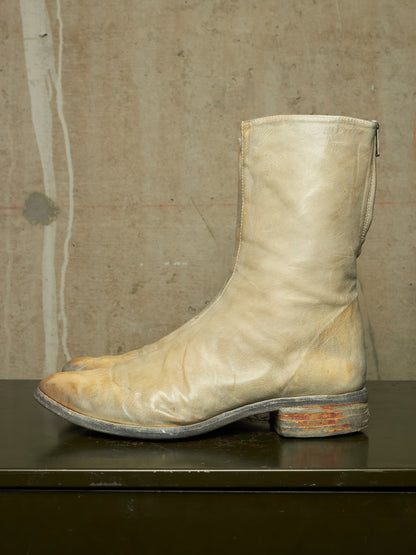 A single light beige leather boot with a distressed, weathered finish, featuring a low stacked heel and a zip closure on the side, resting on a dark surface against a textured concrete wall.