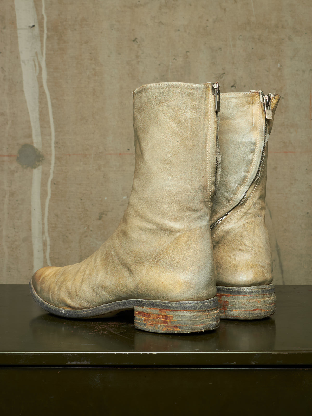 A pair of worn beige leather ankle boots with side zippers, displayed on a dark surface against a textured concrete wall.