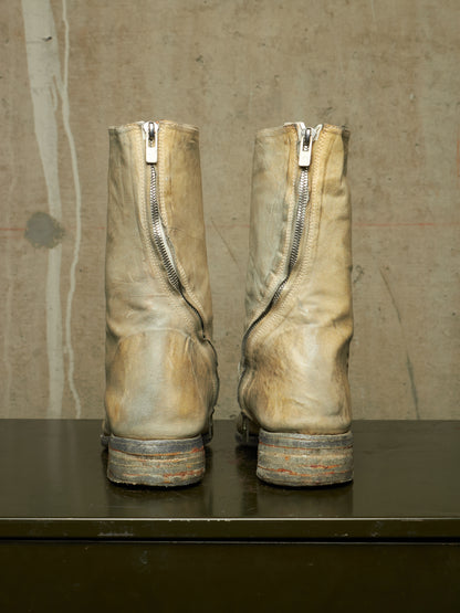 A pair of worn beige leather boots with side zippers, shown from the back on a dark surface against a textured concrete wall.