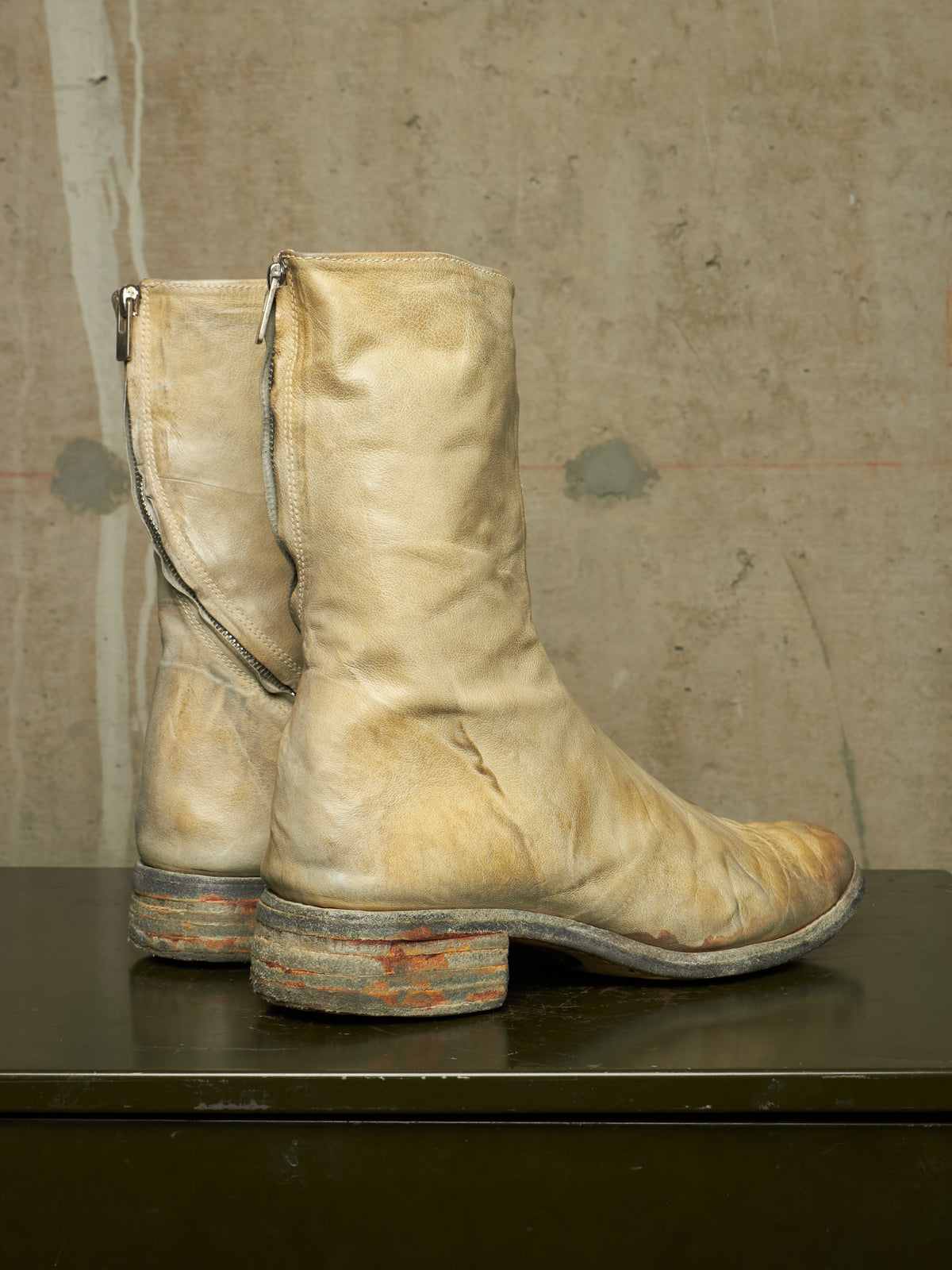 A pair of distressed beige leather mid-calf boots with side zippers, resting on a dark surface against a textured concrete wall.