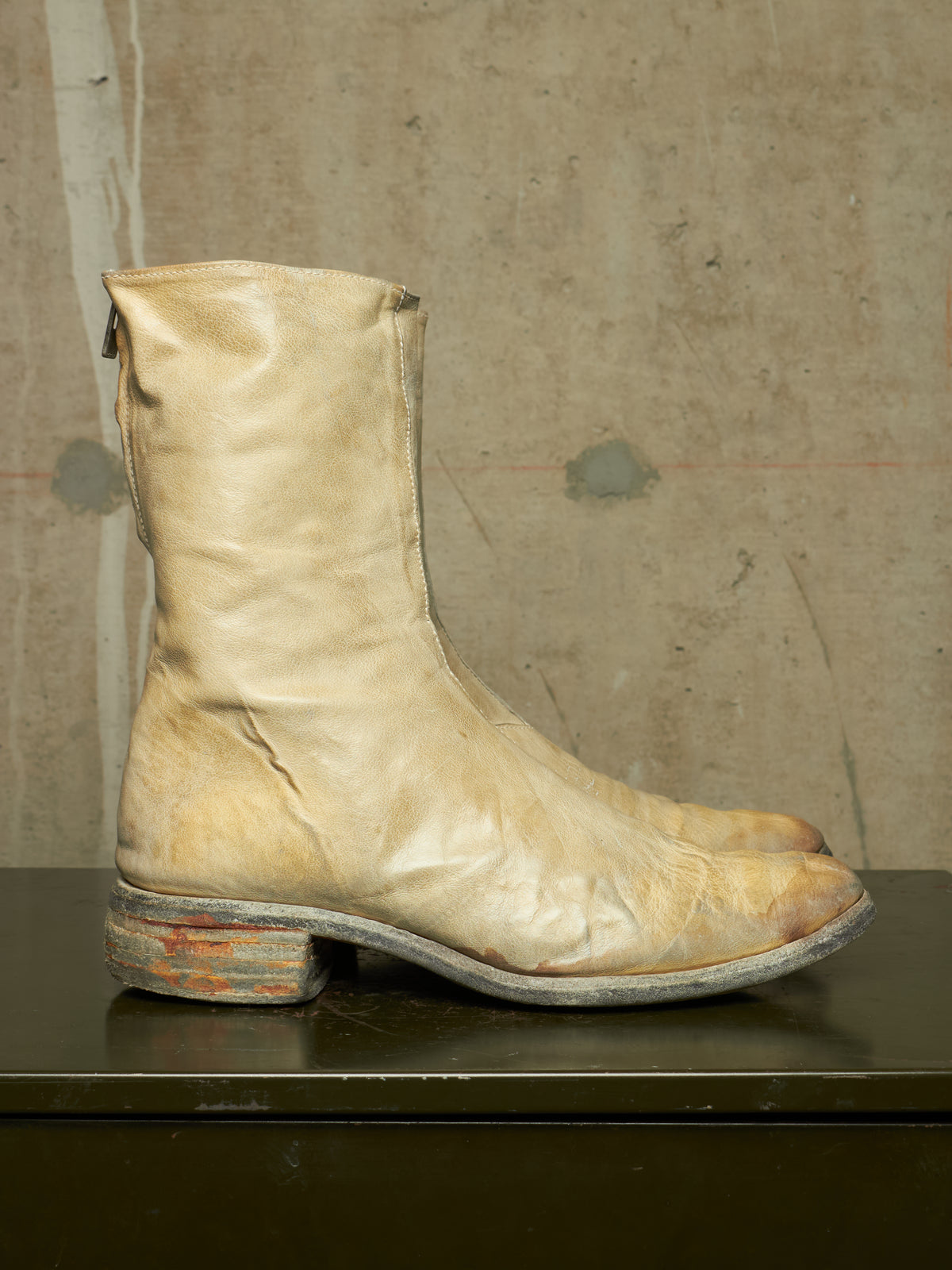 A single light beige leather cowboy boot with a distressed, weathered finish, featuring a stacked wooden heel and a pointed toe, shown against a textured concrete wall.